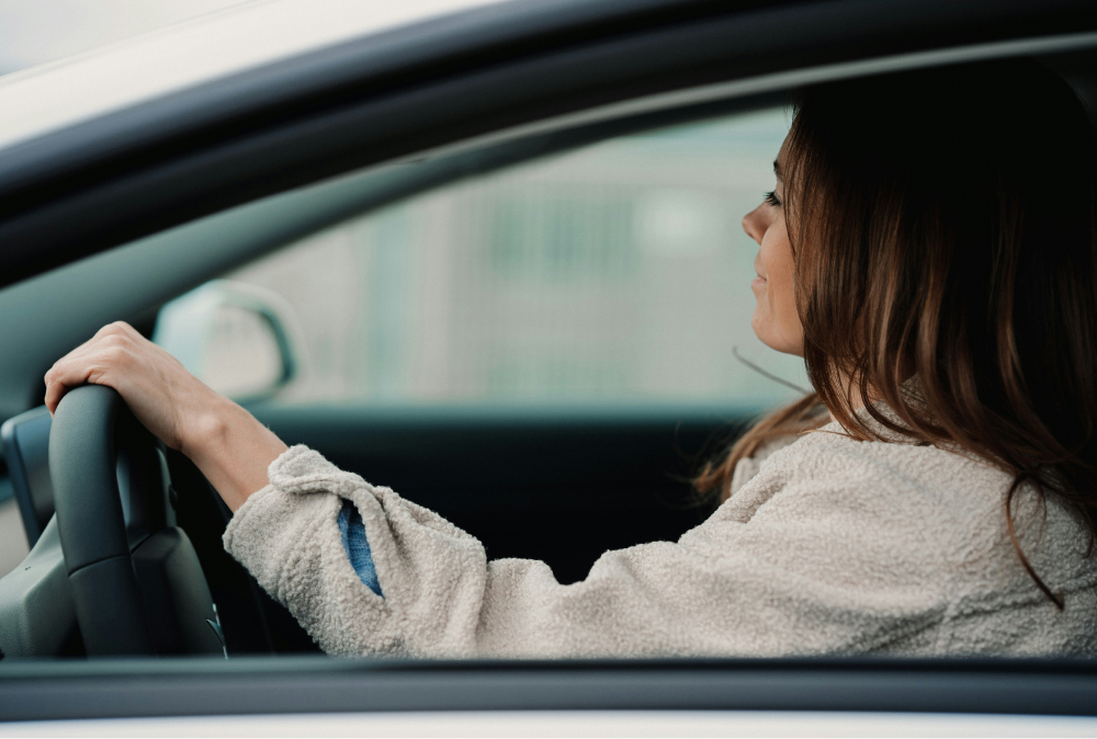 Bild på en kvinna i profil som sitter i en bil med ena handen på ratten